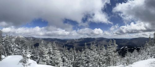 A panoramic view of snow-covered mountains and trees under a partly cloudy sky.
