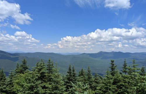 A panoramic view of lush green mountains under a bright blue sky with fluffy white clouds.