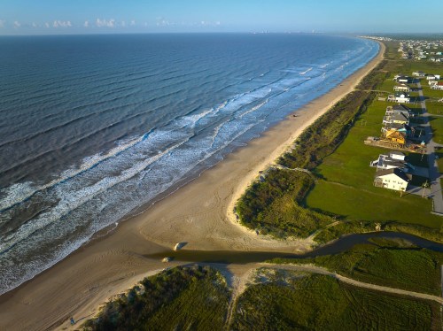Aerial view of a sandy beach with gentle waves, bordered by green grass and houses along the shoreline.