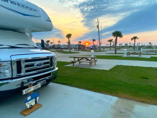 A camper parked at a campsite with picnic tables, palm trees, and a sunset in the background.