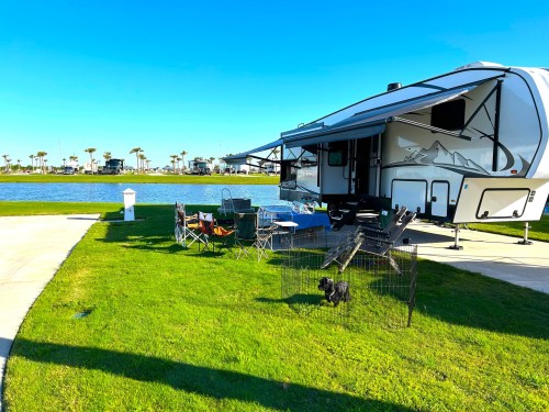 A camper parked by a lake, with chairs set up on the grass and a dog in a pen nearby under a clear blue sky.