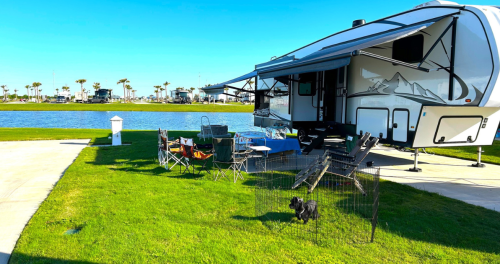 A parked RV by a lake, with outdoor chairs and a small dog in a pen on the grassy area. Clear blue sky in the background.