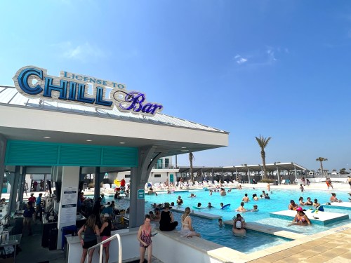 A lively pool scene at Chill Bar, with people enjoying the water and palm trees under a clear blue sky.