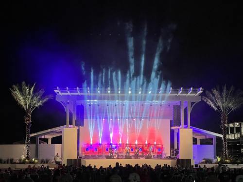 A brightly lit outdoor stage at night, with colorful lights and a crowd gathered in front, palm trees in the background.