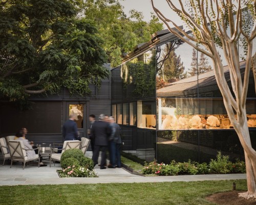 A modern outdoor space with a glass wall, showing people inside a kitchen and guests mingling outside.