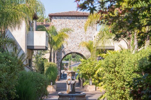 A stone archway surrounded by lush greenery and palm trees, leading to a courtyard with a fountain.