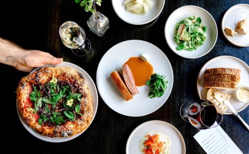 A table set with various dishes, including pizza, salad, and meat, alongside glasses of wine and bread.