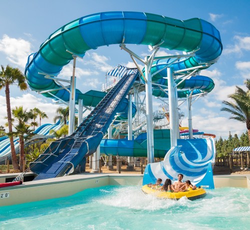 A colorful water slide at a water park with people riding a yellow raft in a splash pool below. Palm trees in the background.