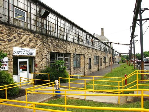 A brick factory building with large windows, a yellow railing, and a sign for a factory outlet. Green grass nearby.