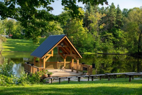 A wooden gazebo by a calm pond, surrounded by lush greenery and trees, reflecting in the water.