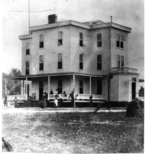 Historic black-and-white photo of a large, multi-story house with people gathered on the porch and lawn.