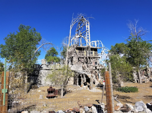 Abandoned structure with twisted metal and trees against a clear blue sky, surrounded by rocky terrain and a fence.