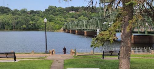 A person stands by a river near a green bridge, surrounded by trees and park benches on a sunny day.