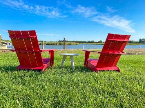 Two red Adirondack chairs facing a calm lake, with a small white table between them, set on green grass under a blue sky.