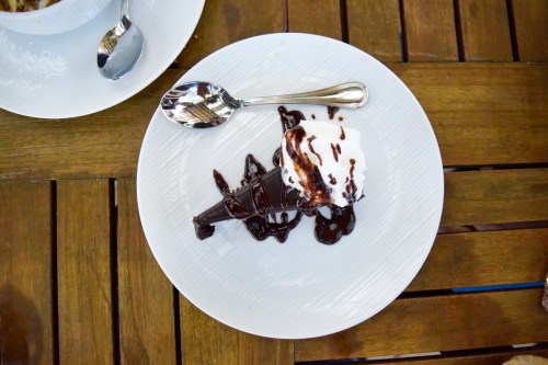 A slice of chocolate cake topped with whipped cream and chocolate drizzle, served on a white plate on a wooden table.