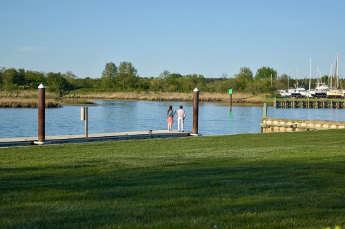 A couple walks hand in hand along a dock by a calm waterway, surrounded by greenery and boats in the distance.