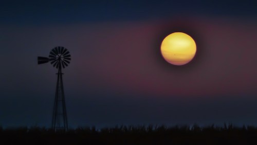 A silhouette of a windmill against a vibrant sunset, with a large sun glowing in the twilight sky.