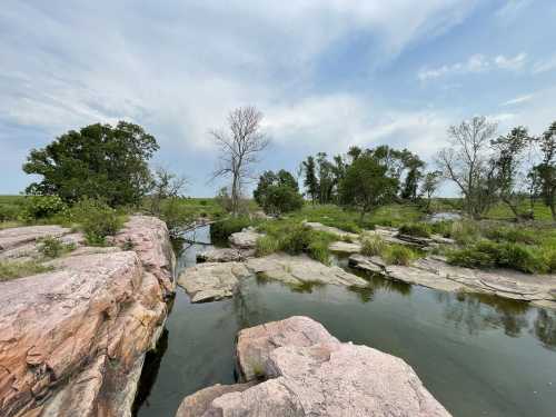 A serene landscape featuring rocky outcrops, a calm waterway, and scattered trees under a cloudy sky.