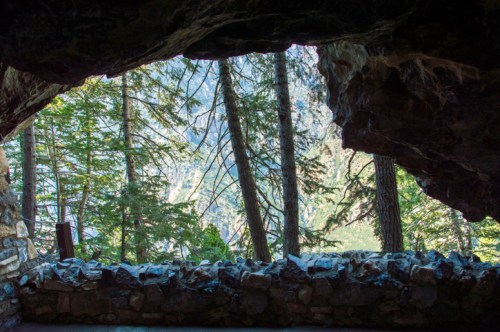 View from a rocky cave opening, framed by trees and mountains in the background. Natural light illuminates the scene.