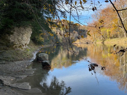 A serene river scene with autumn leaves, rocky banks, and calm water reflecting the trees and sky.
