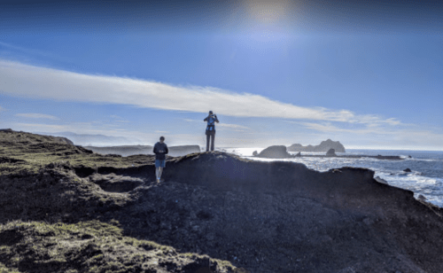 Two people stand on a rocky coastline, with ocean waves and distant cliffs under a bright blue sky.