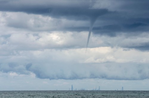 A waterspout forms over the ocean, with a city skyline visible in the background under cloudy skies.