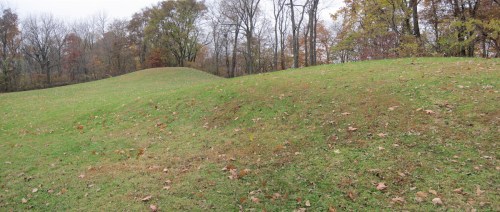 Two grassy mounds in a park, surrounded by trees with autumn leaves, under a cloudy sky.