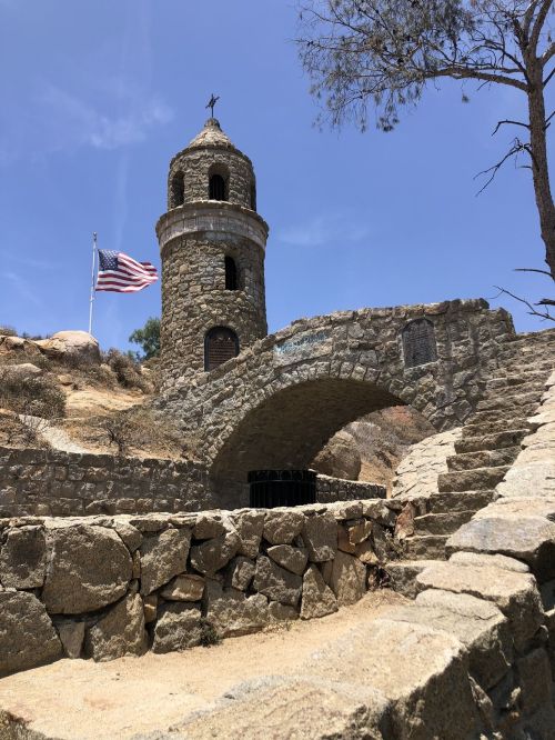 A stone tower with a flag, situated on a hillside with a bridge and steps leading up to it under a clear blue sky.