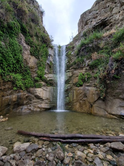 A serene waterfall cascades into a clear pool, surrounded by lush greenery and rocky terrain.