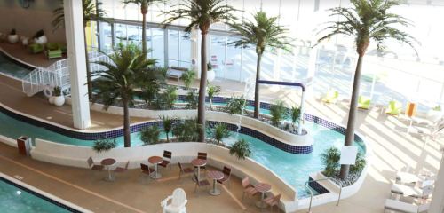 Indoor pool area with palm trees, lounge chairs, and a winding water feature. Bright and inviting atmosphere.