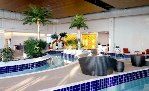 A modern indoor pool area featuring palm trees, lounge seating, and a curved water feature with blue tiles.