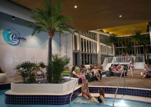 Indoor pool area with palm trees, people relaxing in the water, and a modern design featuring lounge seating.