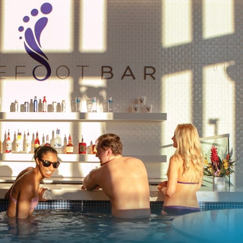 A sunny bar scene with two people enjoying drinks by the pool, surrounded by shelves of colorful beverages.