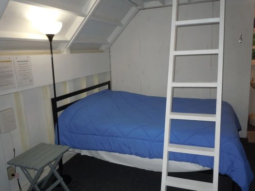 Cozy attic bedroom with a blue quilted bed, a small table, a lamp, and a ladder leading to a loft area.