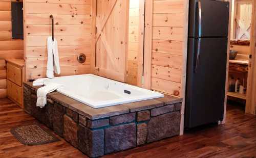 A cozy bathroom featuring a stone-accented jacuzzi, wooden walls, and a refrigerator in a rustic setting.