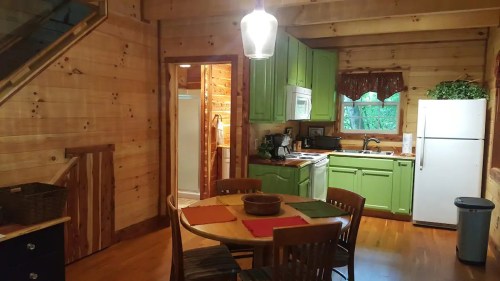 Cozy cabin kitchen with green cabinets, wooden walls, a round dining table, and a view of a bathroom door.