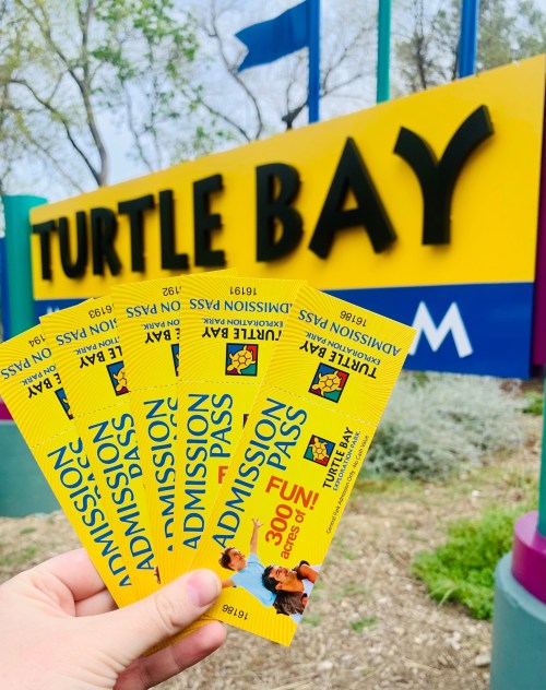 A hand holding four yellow admission passes in front of a colorful Turtle Bay sign.