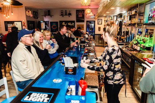A lively bar scene with patrons at the counter and a bartender serving drinks in a cozy atmosphere.