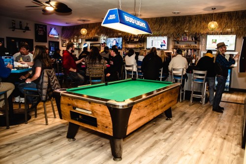 A lively bar scene featuring a green pool table, patrons at the bar, and a tropical-themed decor.