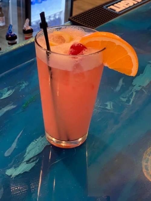 A refreshing pink cocktail garnished with an orange slice and a cherry, served in a tall glass on a blue bar surface.