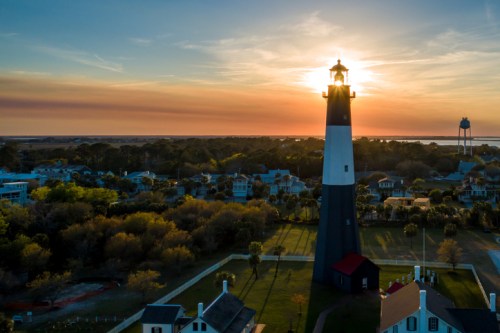 A lighthouse stands tall against a sunset, surrounded by trees and houses, with a water tower in the background.