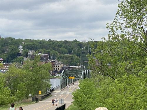 A green bridge spans a river, with people walking and lush trees lining the path under a cloudy sky.