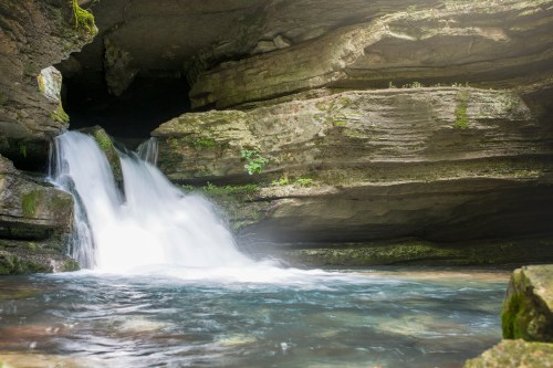 A serene waterfall cascades into a clear pool, surrounded by moss-covered rocks and lush greenery.