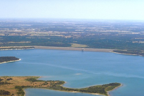Aerial view of a large lake surrounded by green land and a dam in the distance under a clear blue sky.