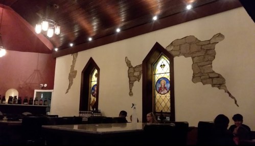 Interior of a restaurant featuring stained glass windows and a rustic stone wall design. Dim lighting creates a cozy atmosphere.