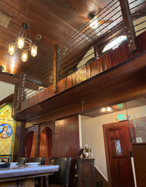 Interior of a rustic space featuring wooden beams, a balcony, and stained glass windows. Warm lighting enhances the ambiance.