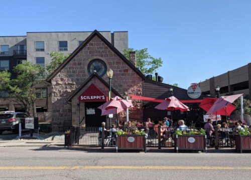 A stone building with a red roof and outdoor seating, featuring umbrellas and flowers, on a sunny day.