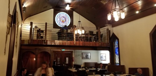 Interior of a restaurant with a high ceiling, stained glass windows, and a loft area with diners.