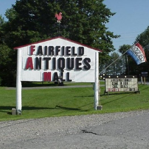 Sign for Fairfield Antiques Mall with a rooster on top, surrounded by green grass and trees.
