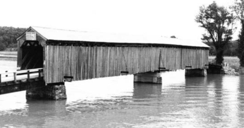 A historic covered bridge spans a river, surrounded by trees, captured in black and white.